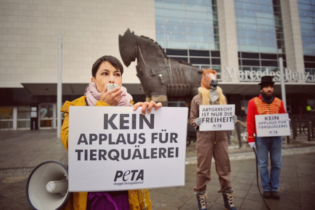 Frau mit Megaphon hält Plakat hoch. Im Hintergrund steht eine Pferdestatue und als Pferd und Dompteur verkleidete Presonen.