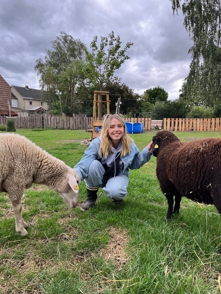 Frau in der Hocke streichelt lächelnd links und rechts von ihr befindende Schafe