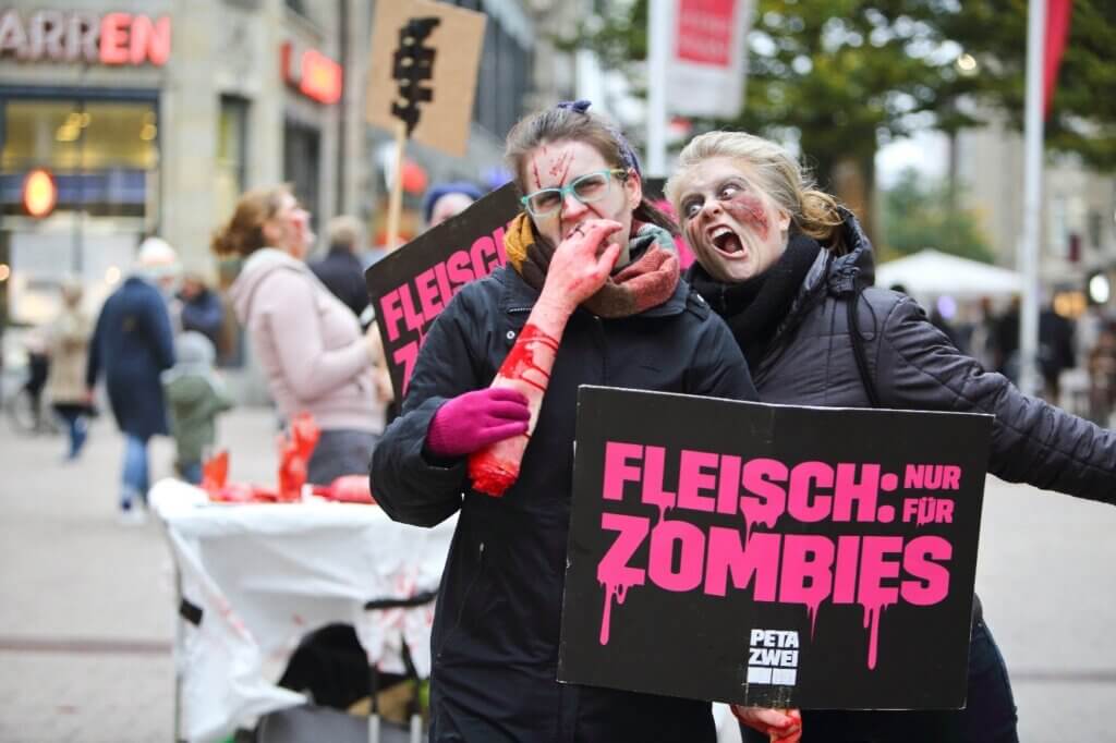Zwei als Zombies verkleidete Personen beißen in einen Arm und halten Plakate