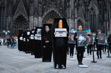 Mehrere Personen mit Tiermasken stehen in hintereinander aufgereihten Särgen vor dem Kölner Dom