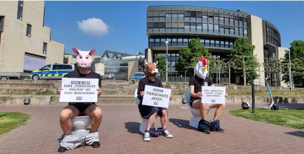 Drei Menschen mit Tiermasken sitzen auf Toiletten vor einem Gebäude und halten Schilder.