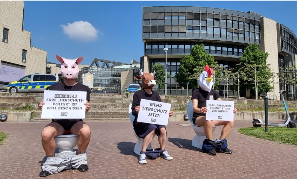 Drei Menschen mit Tiermasken sitzen auf Toiletten vor einem Gebäude und halten Schilder.