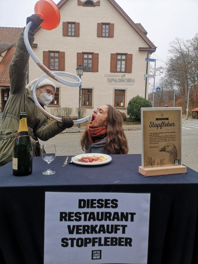 Eine Person kniet hinter einem gedeckten Tisch und wird von einer anderen Person mit einem Trichter gefüttert.