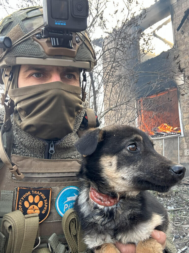 Ein mann in Tarnkleidung mit Helm und Zuch vor dem Mund hält einen Hund auf dem Arm.